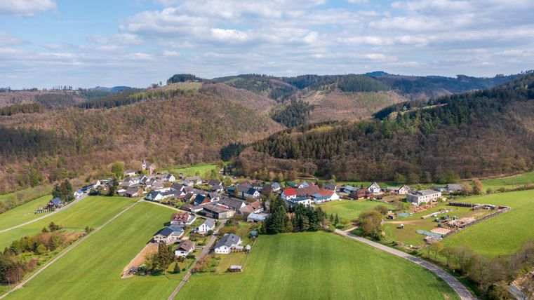 Eine malerische Landschaft mit einem kleinen Dorf umgeben von Hügeln und Wiesen. Im Hintergrund sind bewaldete Berge und ein blauer Himmel zu sehen.