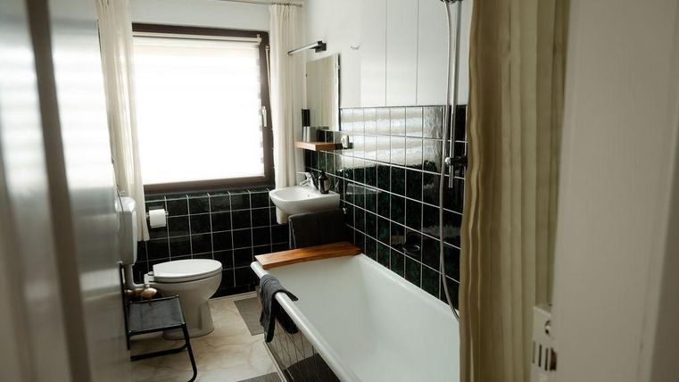 Ein modernes Badezimmer mit einer Badewanne und einer Dusche. Es gibt einen Waschbecken, eine Toilette und große Fenster mit Vorhängen.
