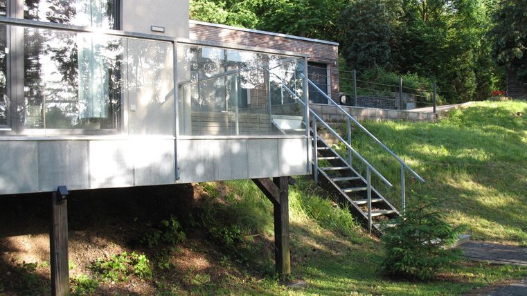 Ein modernes Haus mit einem gläsernen Balkon und einer Treppe. Der Garten ist grün und gut gepflegt.