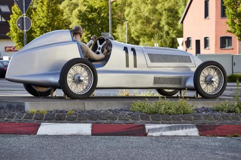 Ein silberner Rennwagen steht auf einer Straße, mit einem Fahrer im Cockpit. Die Umgebung zeigt moderne Gebäude und grüne Bäume im Hintergrund.