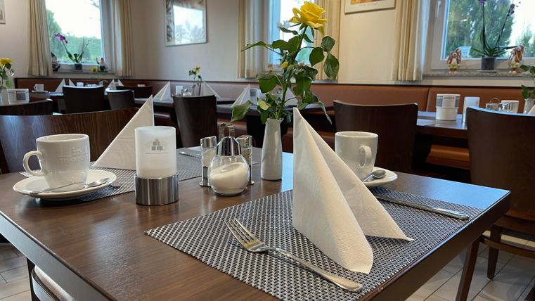 Een uitnodigende tafel in een restaurant met witte servetten, kopjes en een bloemenarrangement. De ruimte is goed verlicht en sfeervol ingericht.