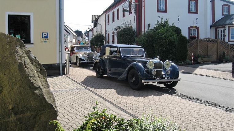 A classic blue car drives through a quiet street in a German village. Traditional buildings and green plants can be seen in the background.