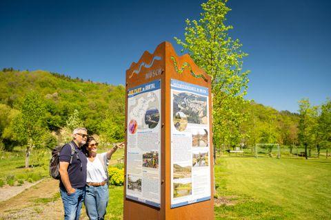 Ein Paar schaut sich einen Informationsstand in einer grünen Parklandschaft an. Die Sonne scheint und die Umgebung ist ruhig und einladend.