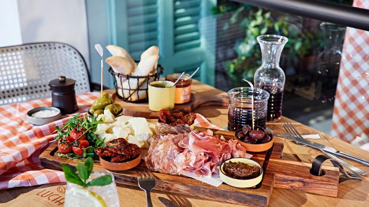 A lovingly arranged board with various types of meat, cheese, and fresh ingredients. There is also bread and glasses with drinks on a wooden table.