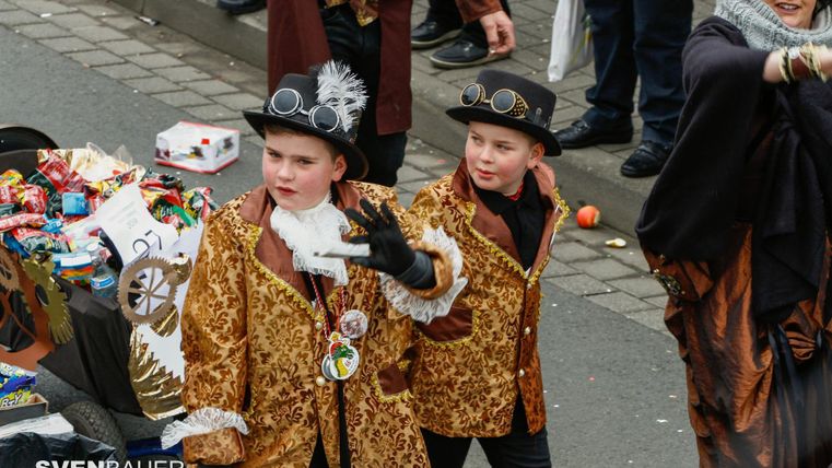 Two children in distinctive costumes with top hats are standing on a street. They are happily posing and enjoying a festive atmosphere.