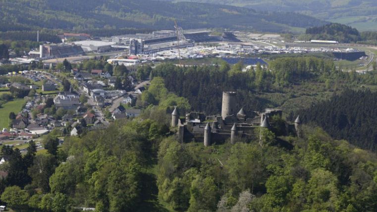 Eine historische Burg ist umgeben von grünen Wäldern und einer kleinen Stadt im Vordergrund. Im Hintergrund sieht man moderne Gebäude und eine Industrieanlage.