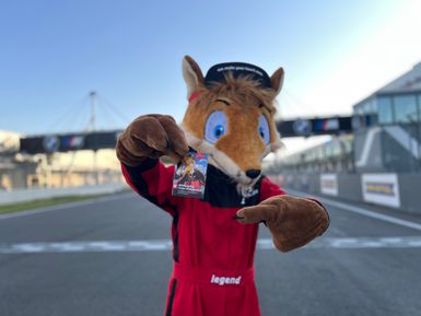 Maskottchen in Form eines Fuchses im roten Rennanzug steht auf der Start-Ziel-Geraden des Nürburgrings und hält einen Erlebnispass in die Kamera. Mit einer Hand zeigt das Maskottchen auf den Pass. Im Hintergrund sind die Rennstrecke, Tribünengebäude und eine BMW-M-Brücke unscharf zu erkennen. Der Fokus liegt auf dem Erlebnispass und dem Maskottchen im Vordergrund.