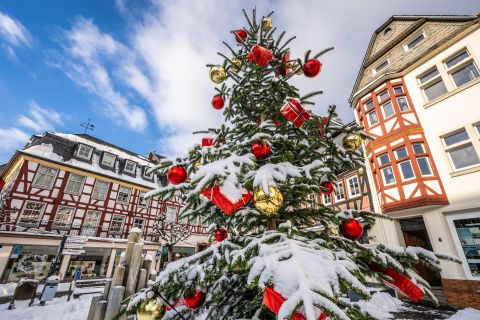 Ein schön dekorierter Weihnachtsbaum ist mit Schnee bedeckt. Im Hintergrund sind traditionell gestaltete Gebäude und ein klarer Himmel zu sehen.