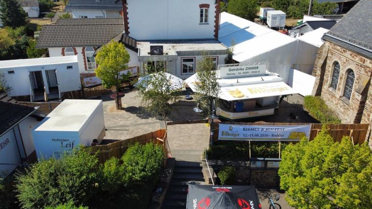 A cozy exterior view of an event venue with trees and booths. There is a large banner announcing an upcoming event.