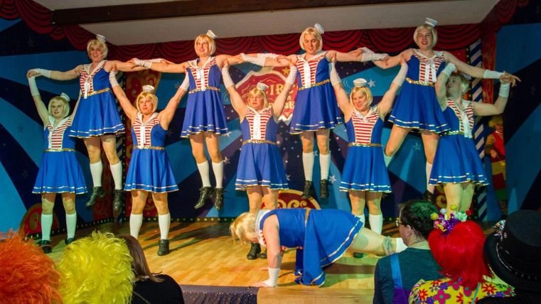 Een groep danseressen in blauwe jurken voert een ingewikkelde choreografie uit. Op de achtergrond zijn blije toeschouwers in kleurrijke kostuums te zien.