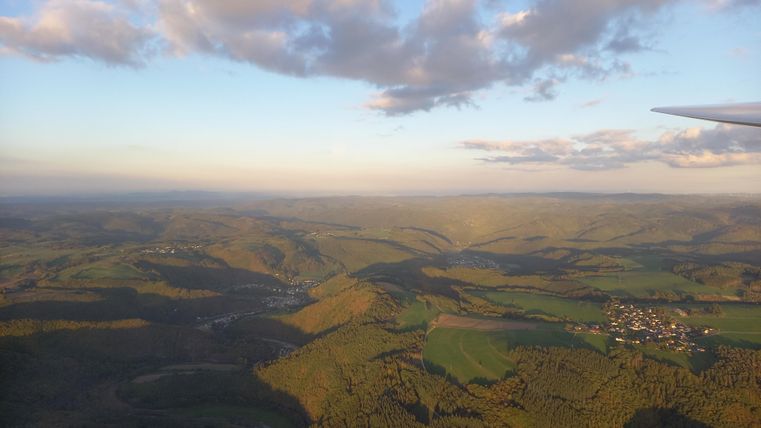 Eine weitläufige Landschaft mit sanften Hügeln und grünen Feldern. Der Himmel ist teilweise bewölkt und das Licht der Abendsonne taucht die Szene in warmes Licht.