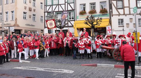 Eine Gruppe von Menschen in roten Kostümen steht auf einer Straße und feiert. Im Hintergrund sind historische Gebäude und eine festliche Atmosphäre zu sehen.