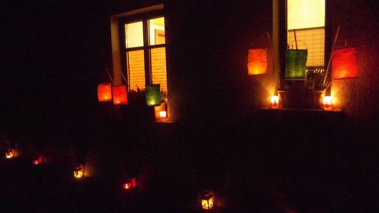 Kleurige lantaarns en kaarsen stralen voor een raam in het donker. Een gezellige en feestelijke sfeer ontstaat.
