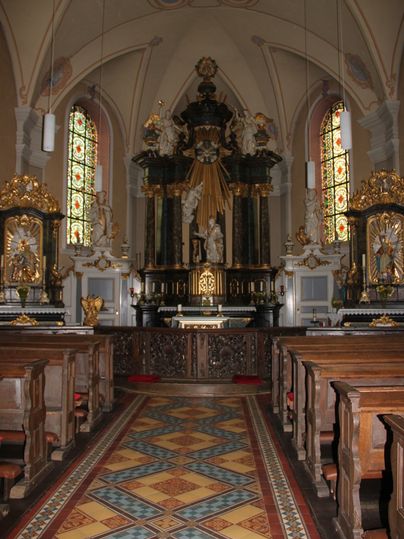 Innenansicht einer Kirche mit Altar, Bänken und bunten Fenstern.
