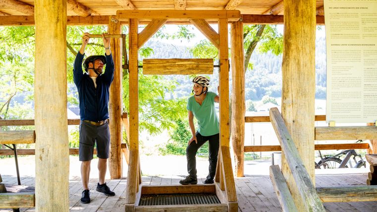 Zwei Personen in Schutzhelmen stehen in einer Holzstruktur. Eine Person zieht an einem Gerät, während die andere zuschaut.