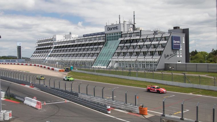A racetrack with multiple cars driving quickly. In the background, a modern building can be seen.