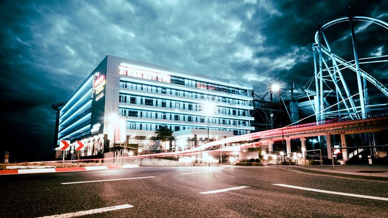 A modern building with bright lights and a dramatic sky. In the foreground, streets are visible with light trails from vehicles.