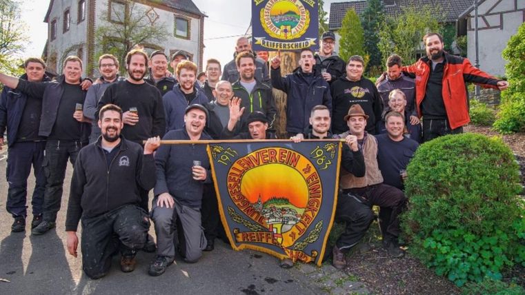 Eine Gruppe von Männern steht lachend zusammen und hält Banner mit einem Sonnenmotiv. Sie tragen einheitliche schwarze Kleidung und befinden sich in einem grünen, ländlichen Umfeld.