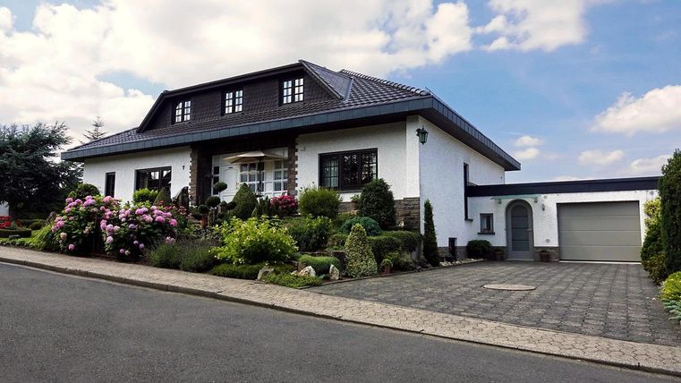 A beautiful house with a well-maintained garden and colorful flowers. The sky is partly cloudy and the street is quiet.