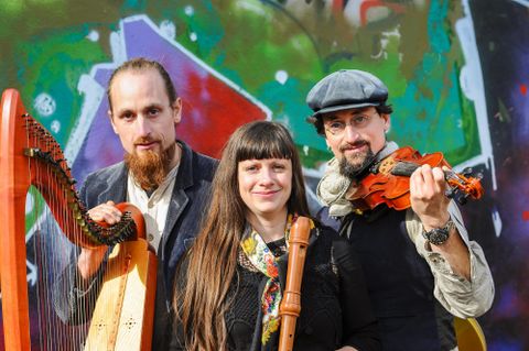 Drei Musiker stehen vor einer bunten Wand. Sie halten verschiedene Instrumente: eine Harfe, eine Flöte und eine Geige.
