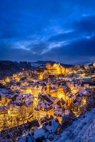 Blick über das verschneite Monschau bei Abenddämmerung: dicht stehende Fachwerkhäuser und historische Gebäude leuchten warm im Kontrast zum tiefblauen Himmel und den weiß bedeckten Dächern.