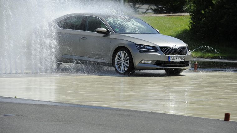 Ein silbernes Auto fährt durch Wasserfontänen auf einem speziellen Testgelände. Die Szene zeigt die Dynamik und Leistungsfähigkeit des Fahrzeugs.
