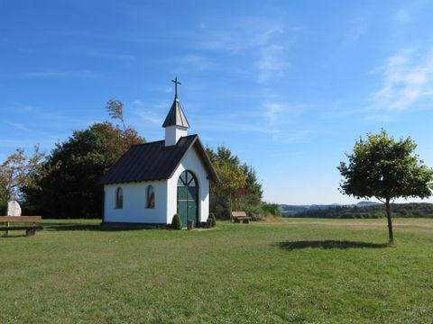 Eine kleine weiße Kapelle mit grünem Tor und Kreuz auf dem Dach steht auf einer Wiese unter blauem Himmel.
