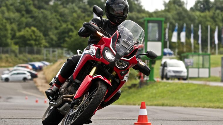 Ein Motorradfahrer auf einem roten Motorrad fährt durch einen Parcours mit Pylonen. Im Hintergrund sind Autos und Fahnen zu sehen.
