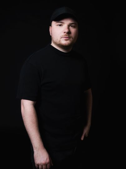 Ein Mann in einem schwarzen T-Shirt und einer schwarzen Kappe steht vor einem dunklen Hintergrund. Er schaut direkt in die Kamera und vermittelt eine ernste Ausstrahlung.