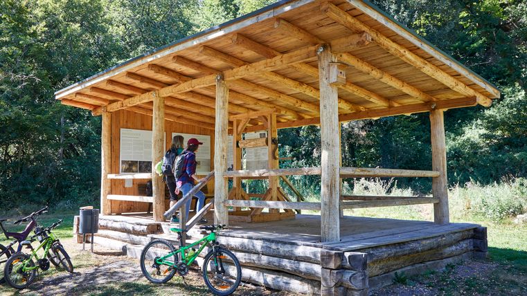 Zwei Personen betrachten Infotafeln in einem hölzernen Unterstand im Freien, umgeben von Fahrrädern und Bäumen.