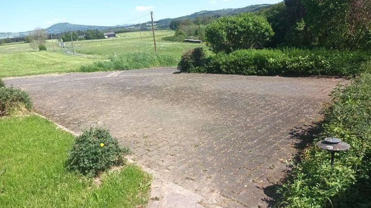 A paved courtyard with green grass and bushes. In the background, fields and hills can be seen.
