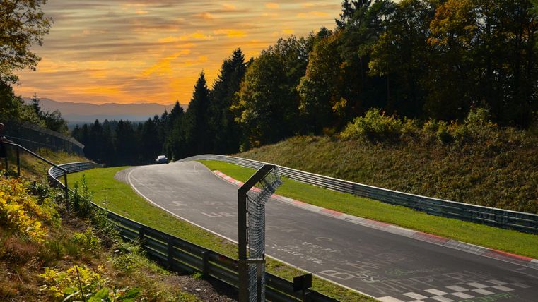 Een bochtige racebaan omringd door bomen en licht heuvelachtig landschap. De lucht is in warme tinten gekleurd tijdens de zonsondergang.