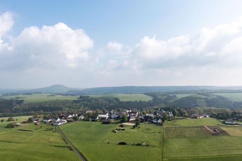 Een schilderachtig landschap met een klein dorp omringd door groene weiden. Op de achtergrond zijn zachte heuvels en een gedeeltelijk bewolkte lucht te zien.
