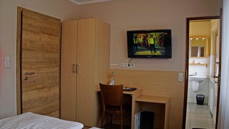 Une chambre simple avec une armoire en bois, une table et une télévision au mur. À travers la porte, une salle de bains est visible.