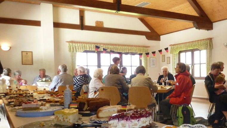 Ein gemütlicher Raum mit vielen Menschen, die an Tischen sitzen und Kuchen essen. Auf den Tischen sind verschiedene Snacks und Süßigkeiten zu sehen.