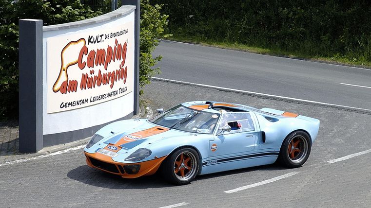 A classic car in blue and orange is standing in front of a campsite sign. The surroundings are green and sunny, perfect for an outing.
