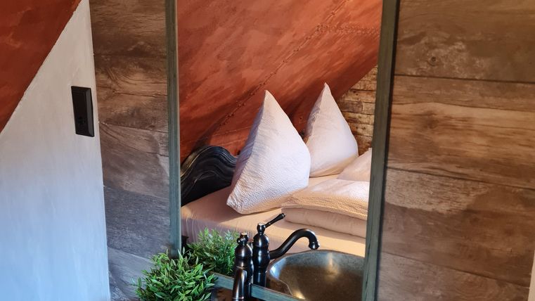 Een moderne wastafel van steen met een grote spiegel en een lamp. Op de achtergrond zijn kussens op een bed te zien.