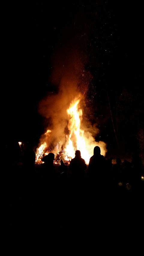Ein großes Feuer brennt in der Nacht, umgeben von dunkler Umgebung. Silhouetten von Menschen sind im Vordergrund zu sehen.