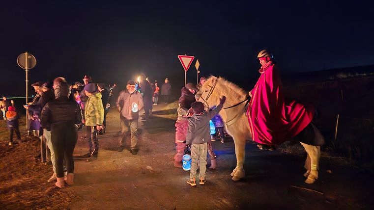 Een groep mensen staat 's nachts langs een straat. Een ridder in een rode mantel rijdt op een paard en praat met de kinderen.