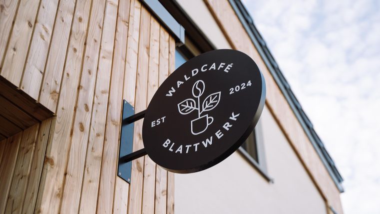 Ein Schild des Waldcafés "Blattwerk" mit einem modernen Design. Das Café ist im Jahr 2024 gegründet worden und zeigt eine natürliche, einladende Atmosphäre.