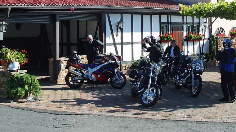 Een groep motorrijders staat voor een traditioneel gebouw met bloeiende planten. Meerdere motoren staan op het geplaveide plein.