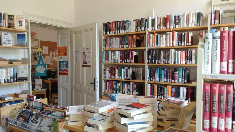 Ein gemütlicher Raum mit Regalen voller Bücher für Kinder. Auf Tischen stehen bunte Spielsachen und kreative Materialien.