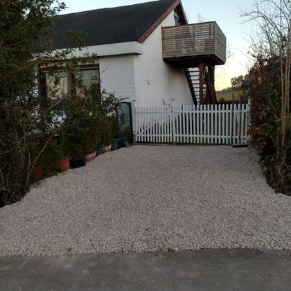 A driveway with gravel and a white fence. In the background, there is a house with a balcony.