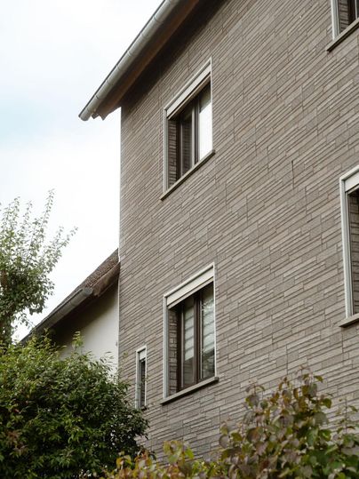 Eine moderne Wohngebäudeansicht mit grauer Fassade und mehreren Fenstern. Grüne Pflanzen umgeben das Gebäude und verleihen der Szene ein gemütliches Ambiente.