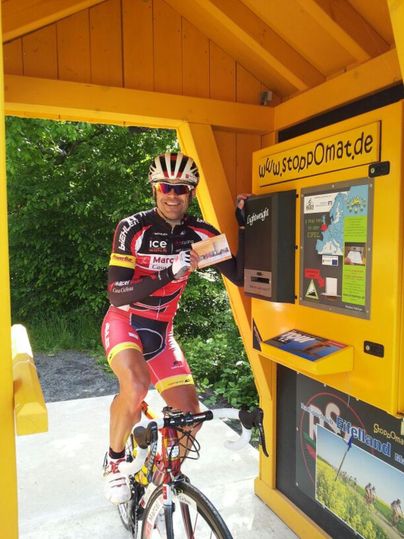 Ein Radfahrer steht neben einem gelben Automaten in der Natur. Er zeigt glücklich eine Karte und trägt sportliche Radkleidung.