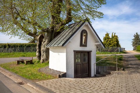 Ein charmantes kleines Häuschen steht an einer Straßenecke, umgeben von Bäumen und Grün. Der blaue Himmel und die ruhige Umgebung verleihen dem Bild eine friedliche Atmosphäre.