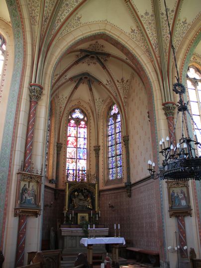 Ein wunderschöner Kircheninnenraum mit hohen gotischen Bögen und bunten Fenstern. Die Wände sind kunstvoll verziert und der Altar ist gut sichtbar.