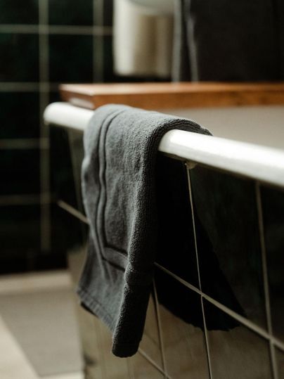 Ein schwarzes Handtuch hängt an der Kante einer Badewanne. Im Hintergrund sind grüne Fliesen und ein Holztisch sichtbar.