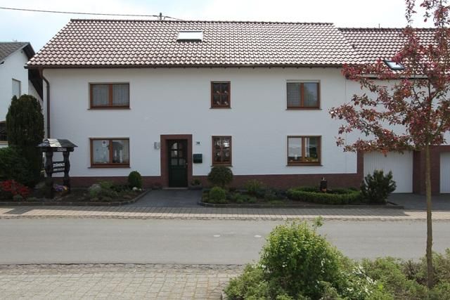 Een twee-verdiepingen tellend, wit woonhuis met een rood pannendak. Voor het huis bevindt zich een verzorgde tuin en een kleine oprit.