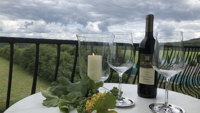 Une table sur un balcon avec deux verres à vin, une bouteille de vin et une bougie. La vue montre des champs verts sous un ciel nuageux.